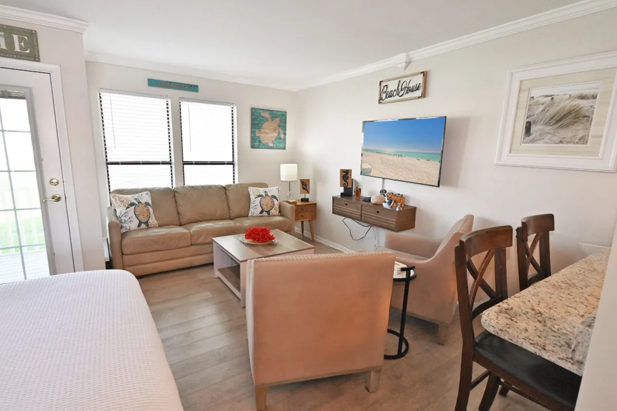 A wide view of the studio's living area, featuring a plush sofa, a smart TV, and a dining table set against large windows. The room is bright, stylish, and filled with natural light, emphasizing coastal-inspired décor and modern finishes.