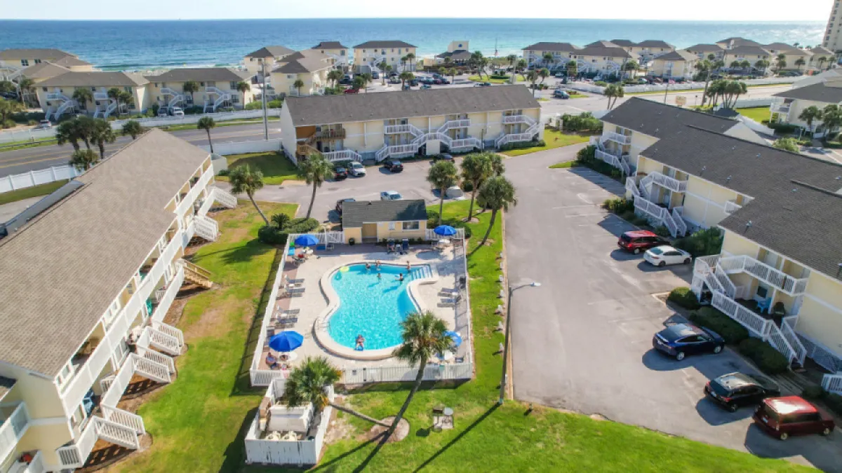 A sparkling swimming pool is at the center of this lively scene, surrounded by resort buildings and offering an inviting ocean view in the background. Guests can be imagined sunbathing, swimming, or socializing by the pool, capturing the essence of shared resort amenities available to residents and guests.
