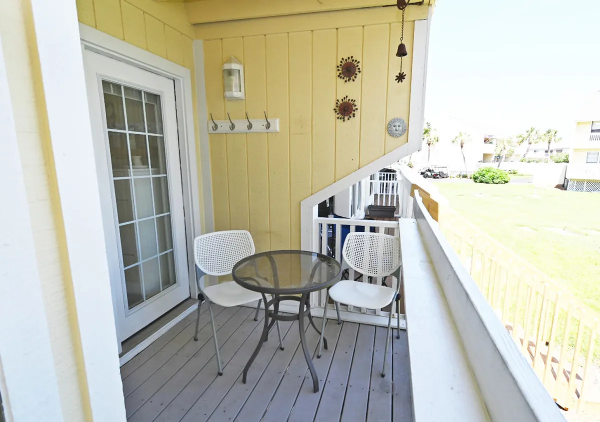 A cozy patio table and two chairs set against a sunny yellow wall adorned with a decorative clock and cheerful sun motifs. The setting suggests a spot for morning coffee, evening relaxation, or casual outdoor meals, all within arm’s reach of the beach.
