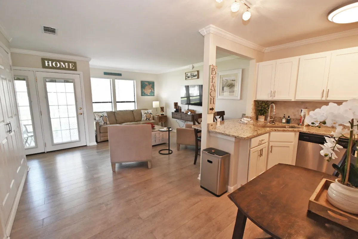 The main living area with a sofa, kitchen island, dining table, and decorative signage. Morning light streams through the windows, highlighting the room’s welcoming ambiance and flexible layout suitable for gatherings or quiet moments.