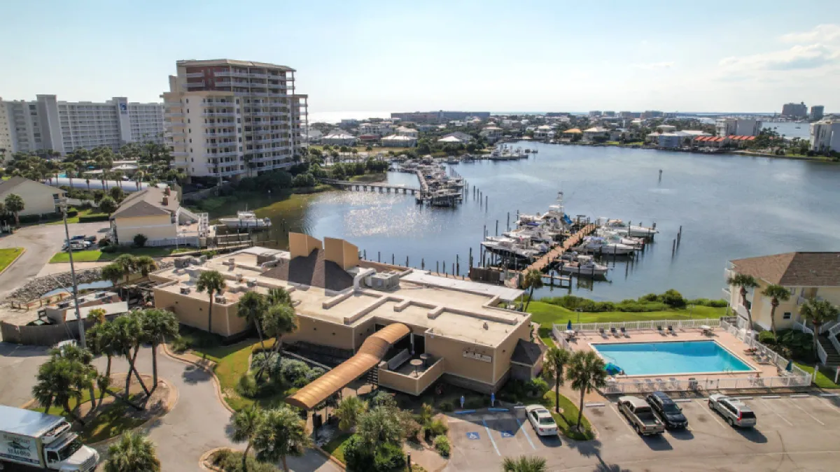 A panoramic scene encompassing the marina, a swimming pool, landscaped gardens, and residential buildings. People are seen boating, swimming, and gathering, with the lush resort grounds and waterfront at their doorstep.