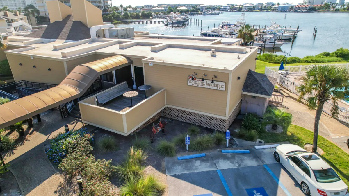 A stylish restaurant building sits beside an outdoor seating area overlooking a tranquil water view, framed by palm trees. Guests dine alfresco, savoring local cuisine and the scenic backdrop.