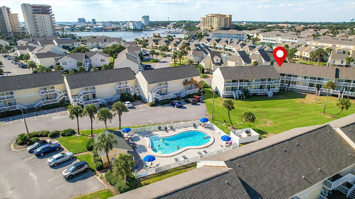 A vibrant outdoor swimming pool is surrounded by palm trees and adjacent residential buildings, with people gathering to swim, sunbathe, and socialize. The setting is both inviting and relaxing, suggesting a lively community atmosphere.