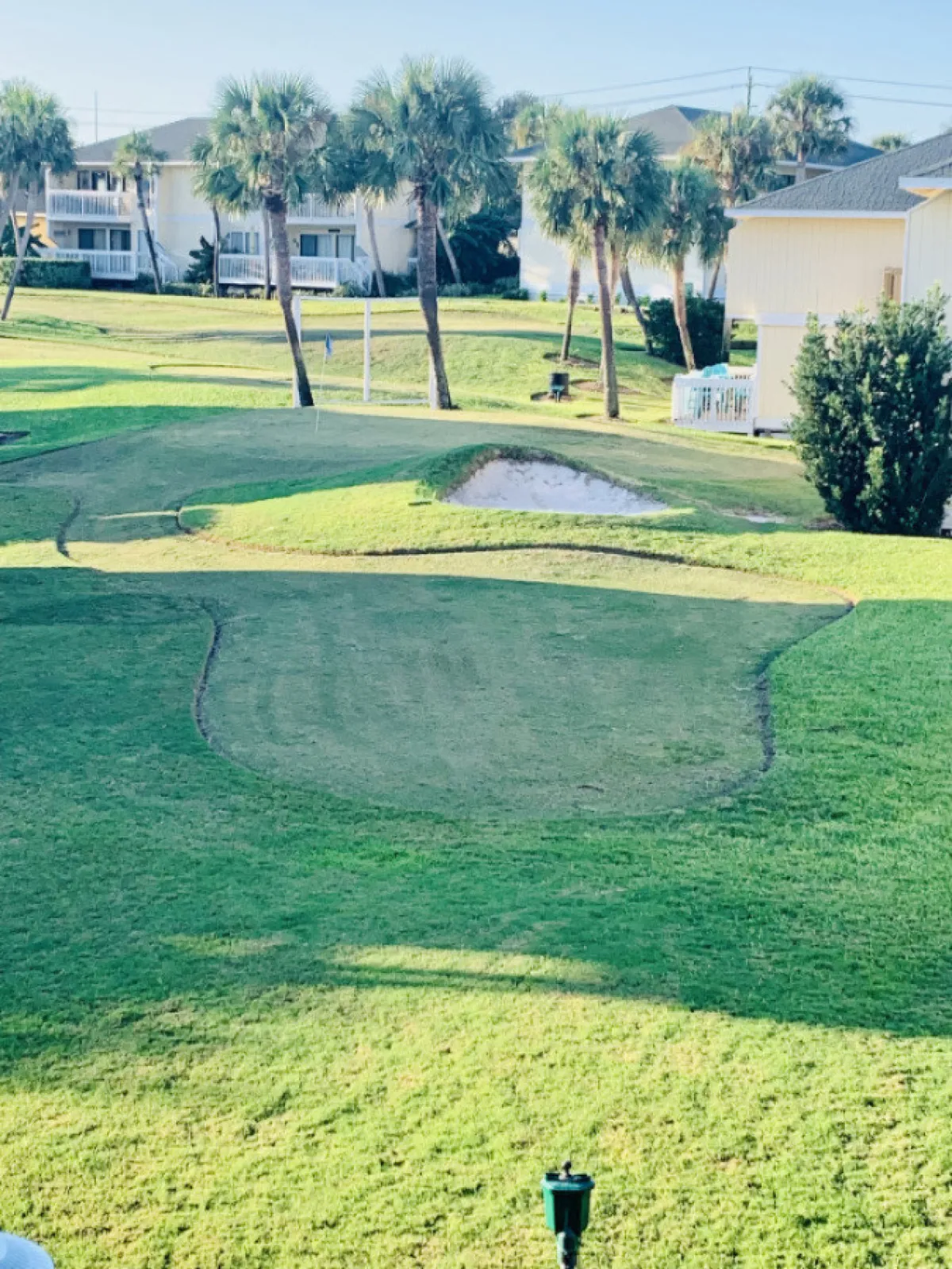 A lush golf green with palm trees and a sand trap, surrounded by houses, captures a leisurely afternoon scene. The area is calm, with people poised to play or enjoy the scenery, evoking the spirit of resort recreation.