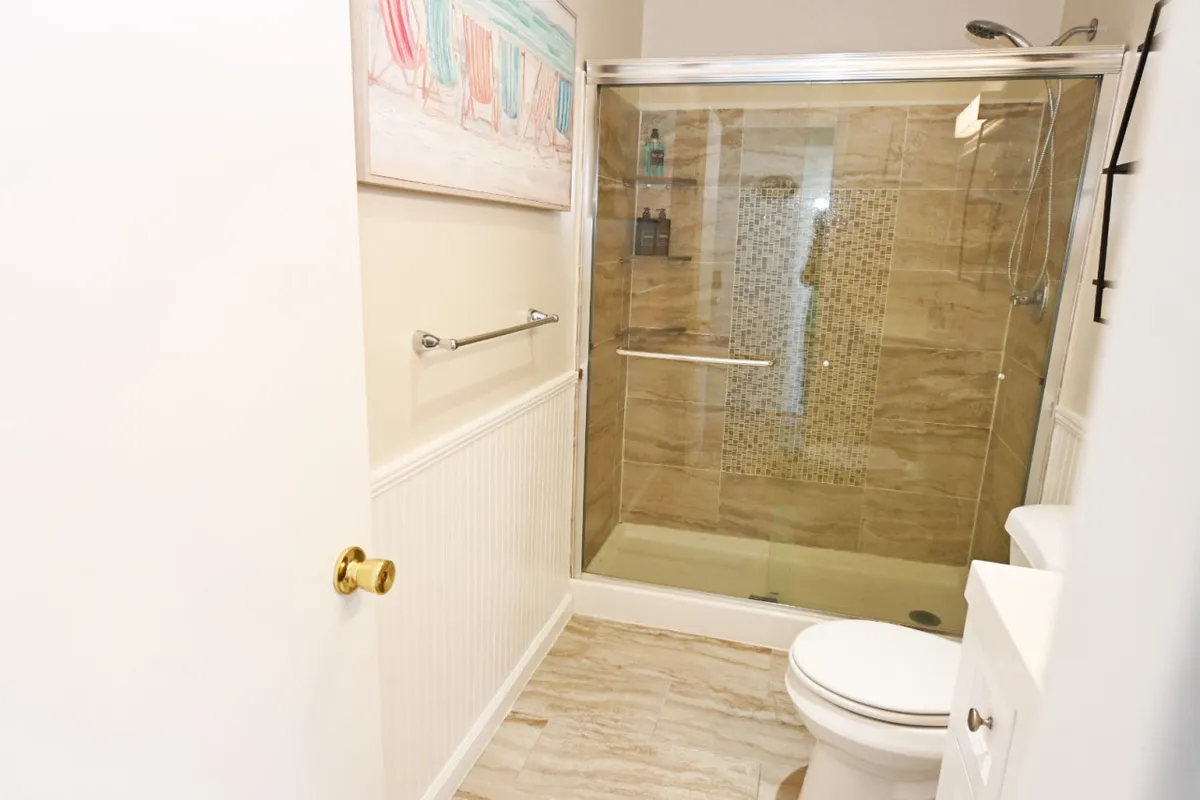 This inviting bathroom scene features a gleaming glass shower enclosure framed by elegant, textured tiles. Light-colored cabinetry and beach-themed artwork add a touch of style, while the soft natural light streaming in enhances the serene ambiance. The attention to detail suggests a space designed for rejuvenation and indulgence.