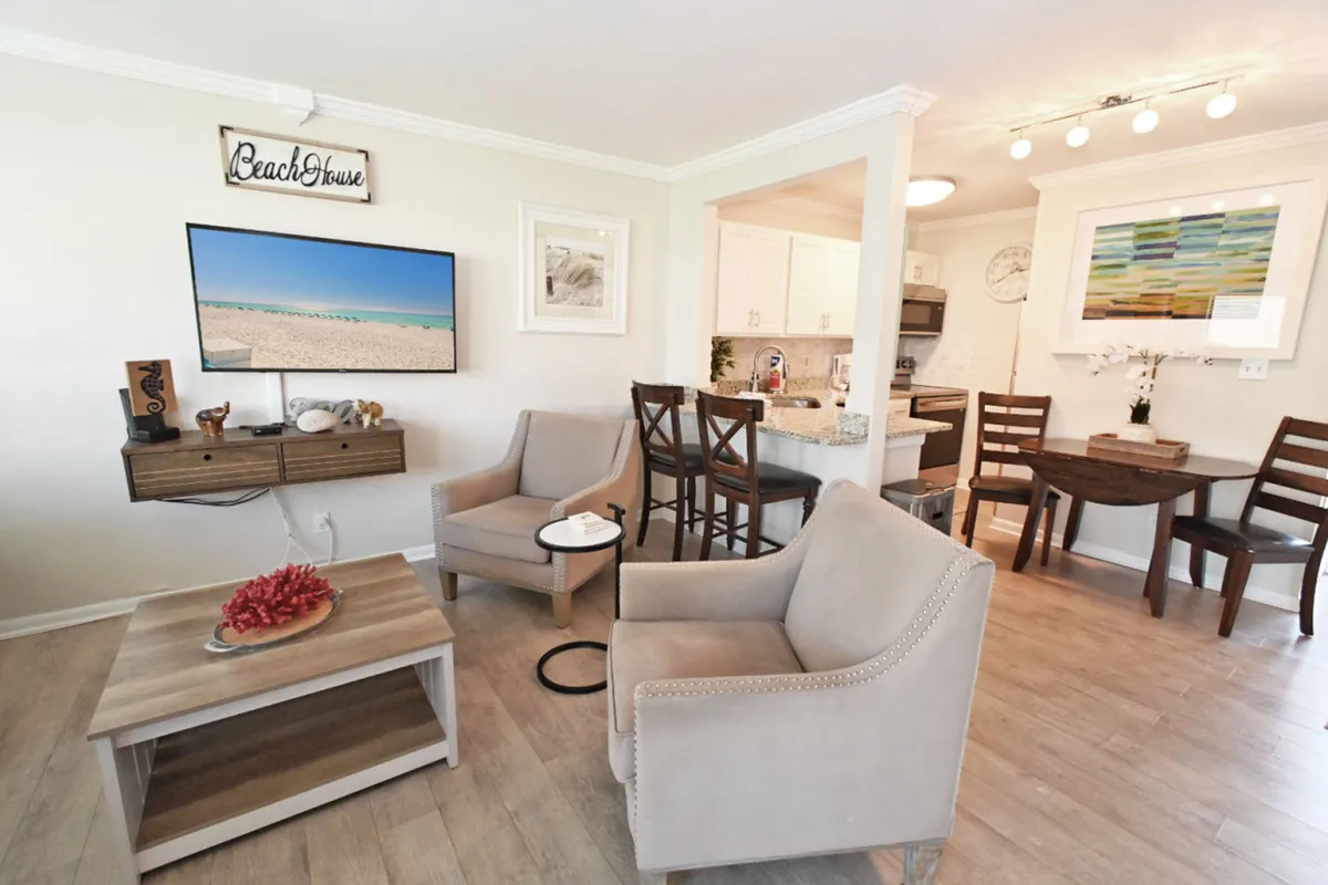 The image showcases a bright, inviting living space with comfortable beige armchairs facing a flat-screen TV displaying a serene beach scene. A wooden coffee table with a coral centerpiece adds coastal charm, while a kitchen counter with bar stools creates a space for casual dining or conversation. The overall atmosphere is relaxed and beach-inspired, perfect for unwinding after a day in Destin.