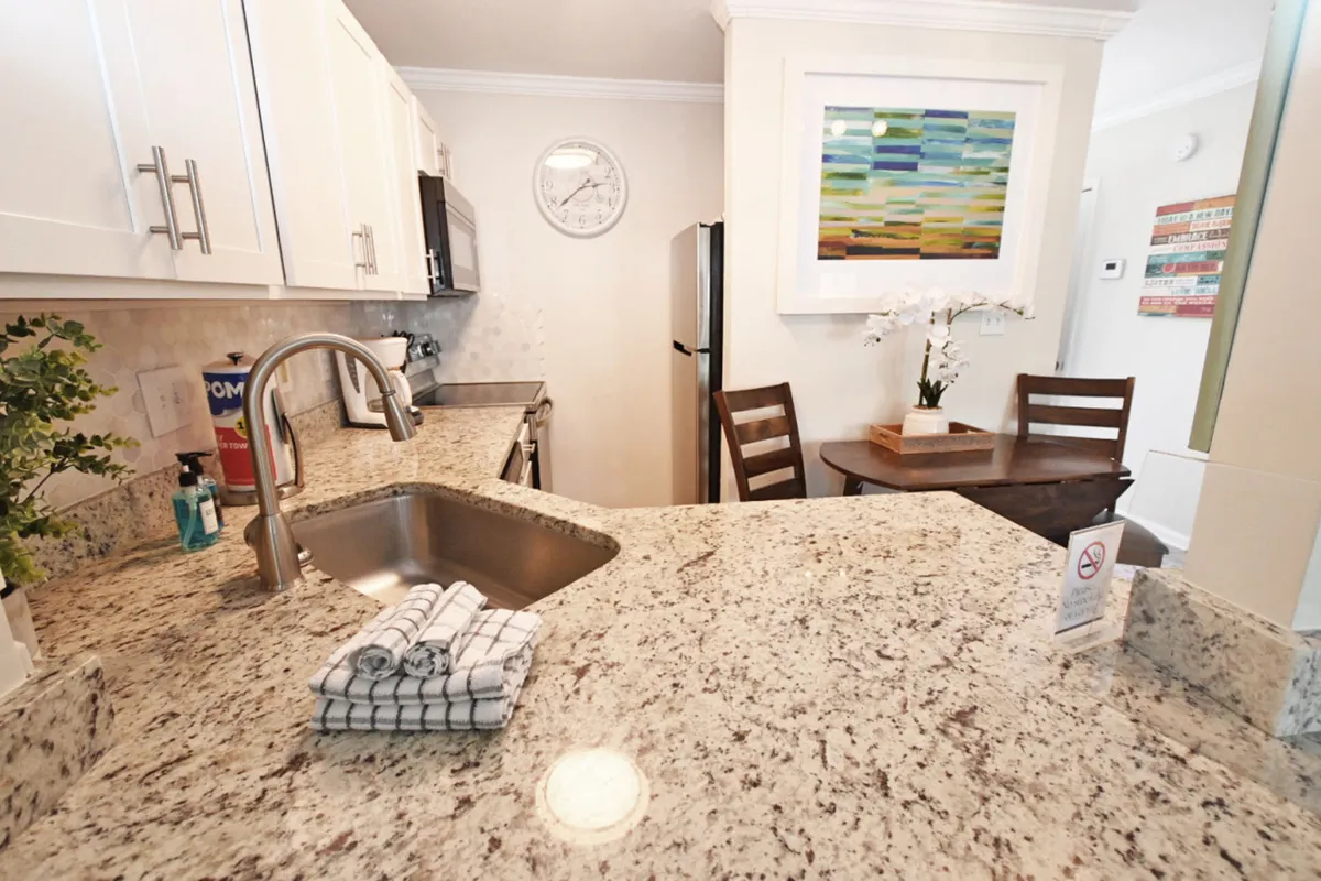 A view of the granite kitchen countertop with sink and neatly folded dish towels, a small round dining table with two chairs, modern appliances (stove and refrigerator), and vibrant wall art above the dining area. The scene is bright and relaxed, emphasizing the kitchen's functionality and inviting feel.