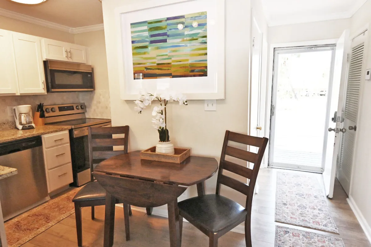 A warm, casual dining space with a small wooden table set for three, modern kitchen appliances including a stove and dishwasher, and a white orchid centerpiece. The colorful abstract wall art adds a touch of beachside calm and the view through the door suggests a seamless transition to outdoor relaxation.