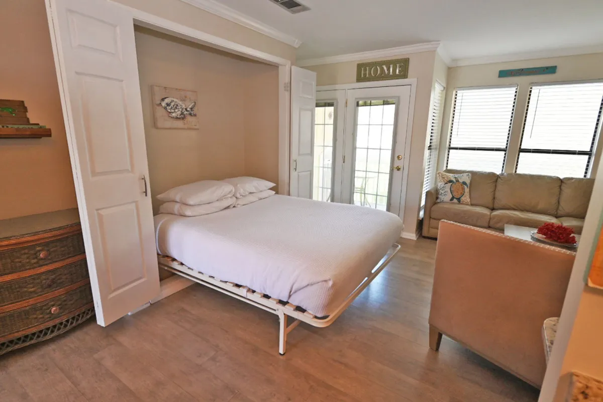 This image features the main living/sleeping area with the Murphy bed folded down, a cozy sofa set near sunlit windows, and French doors leading to a balcony or patio. The decor is beach-themed, including a 'HOME' sign and shell artwork, embodying a relaxed beach cottage feel.