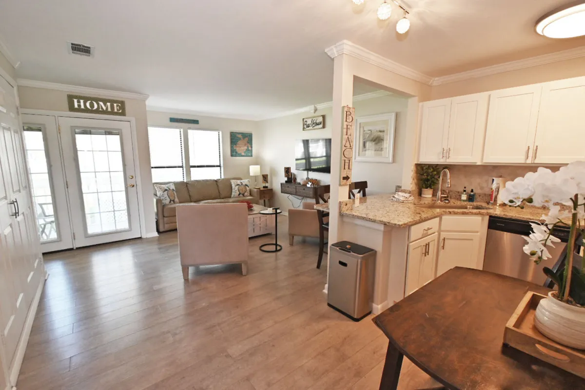This image focuses on the open-plan kitchen and living area, with a comfortable sofa and armchair, flat-screen smart TV mounted on the wall, granite countertop, and decorative beach-themed signs and artwork. The mood is inviting and cozy, ideal for unwinding after beach adventures.