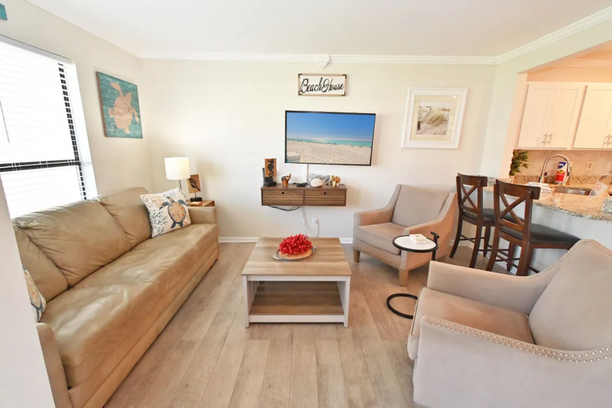 A cozy interior space featuring a tan leather sofa, beach-themed wall art, and a flat-screen TV creates a welcoming atmosphere. The granite kitchen countertop and bar stools suggest a space for preparing and sharing meals, echoing the home-like touches described in the section.