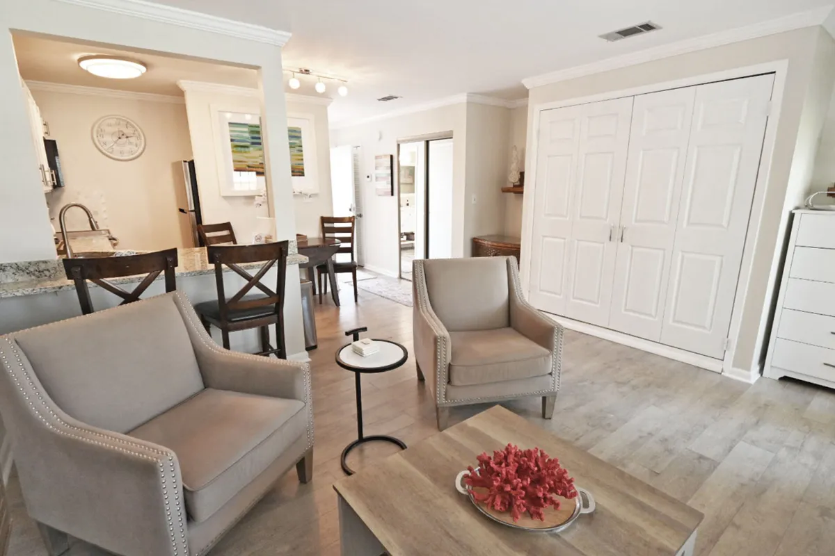 This image shows a calm, relaxed living space with gray armchairs, a wooden coffee table featuring a coral centerpiece, a dining table with chairs near the kitchen, and closed Murphy bed closet doors. The decor enhances the coastal vibe, and the space is designed for both intimate conversations and shared meals or games.