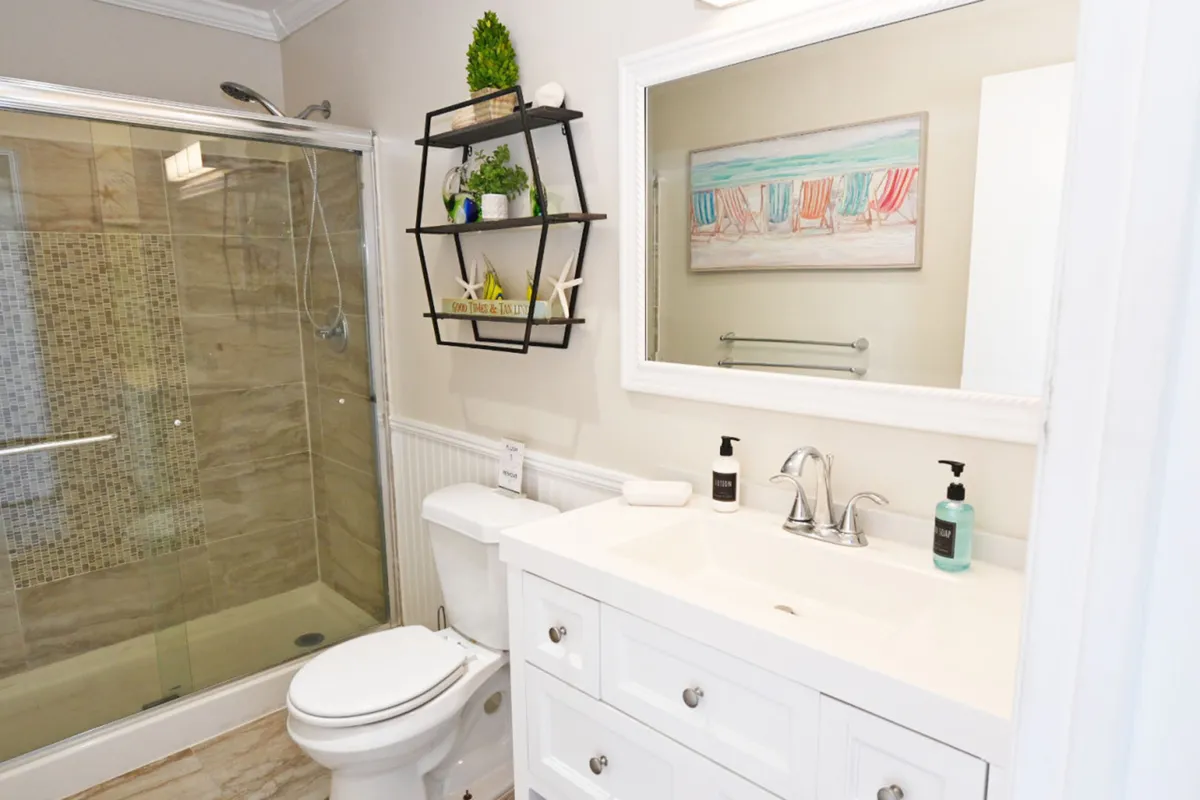 A modern bathroom featuring a glass-enclosed shower with stylish mosaic tile accents. The white vanity with sink and large framed mirror enhances the sense of space and cleanliness, while a decorative wall shelf with plants and beach-themed items adds a thoughtful, welcoming touch.