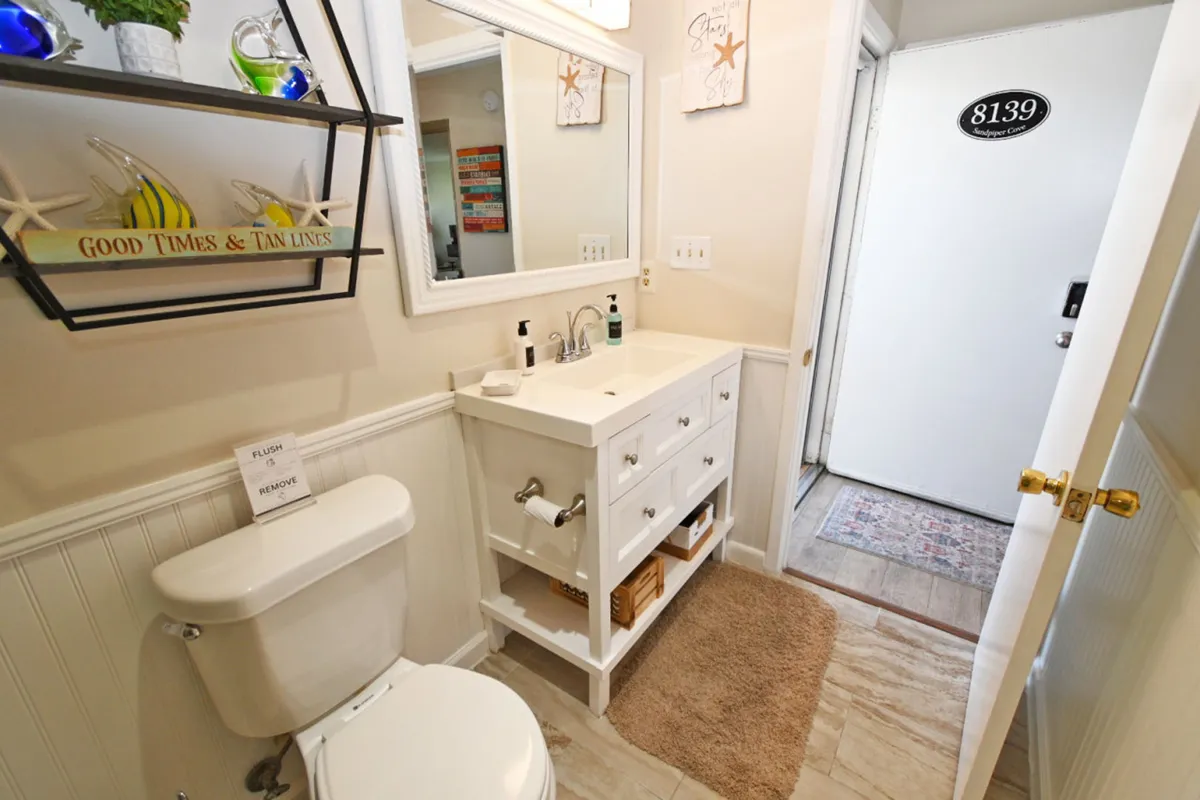 A close-up of the bathroom vanity, featuring a white sink and mirror, decorative shelf with beach-themed items, and a glimpse of the entrance to the unit. The space looks clean and well-stocked, highlighting the availability of essential bath products and amenities.