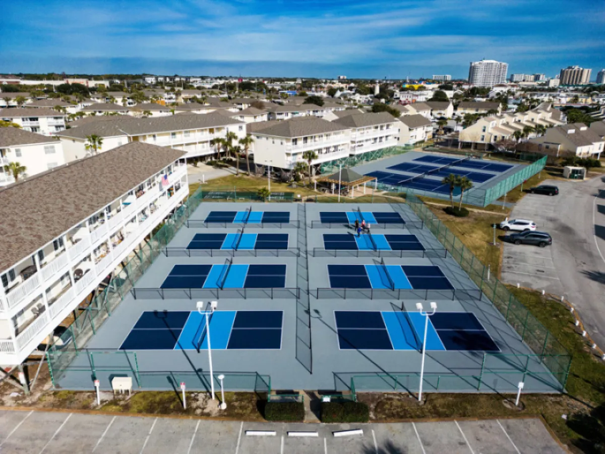 A wide exterior shot of eight pickleball courts with blue and gray surfaces, adjacent residential buildings, and a few players enjoying a game. The surrounding parking lot and landscaped areas reinforce the active, community-focused resort environment.