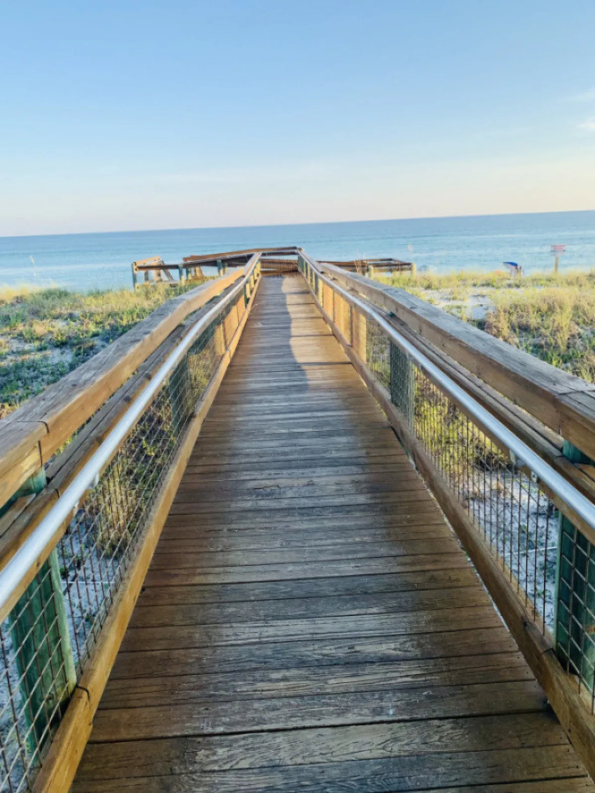 A peaceful wooden boardwalk flanked by sandy dune grasses leads directly to the expansive, calm ocean horizon. The scene captures the serene moment when visitors transition from the community toward the pristine beach, with the endless horizon of the Gulf of Mexico inviting relaxation and exploration.
