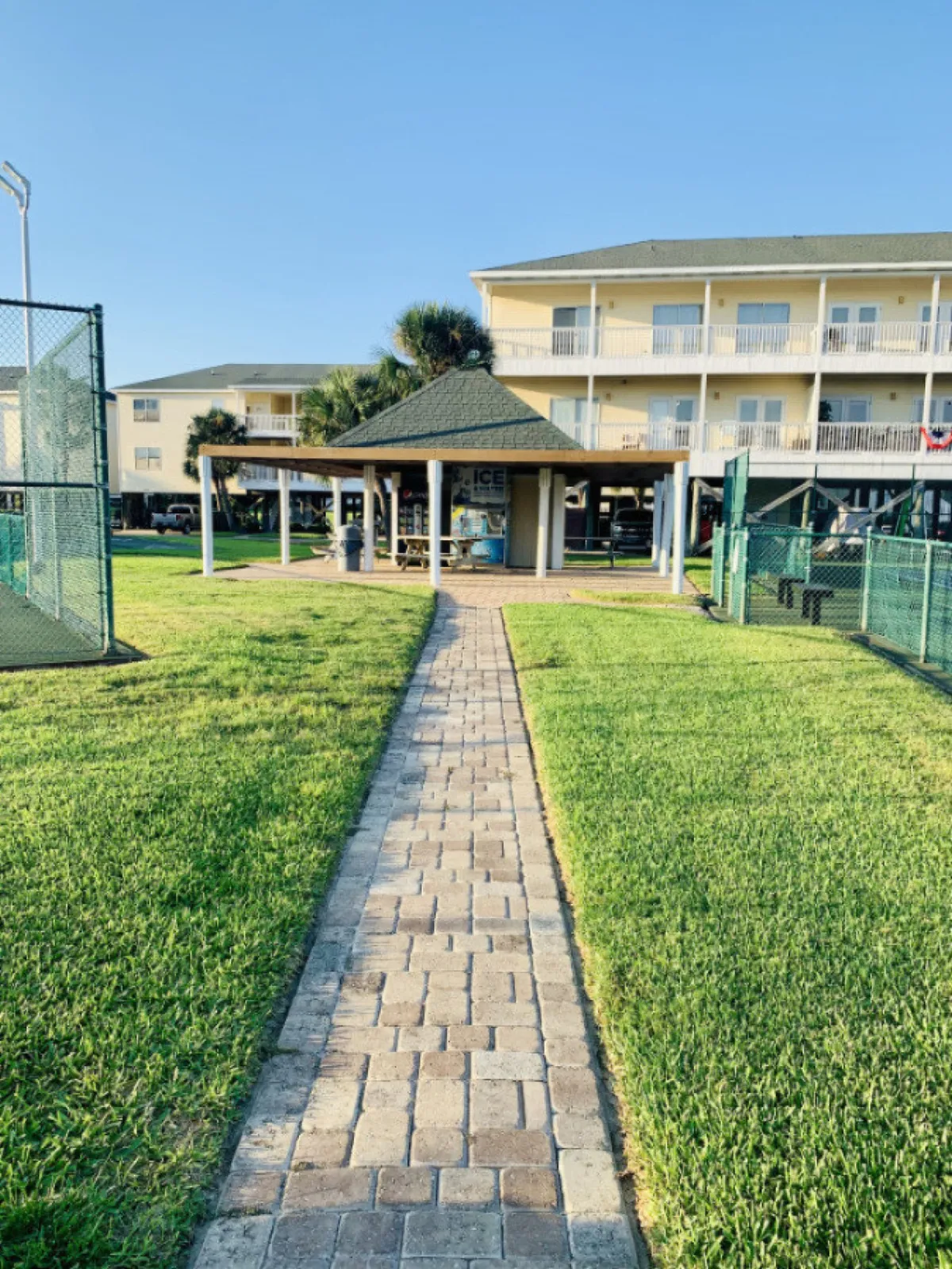 A paved walkway leads to a covered pavilion with seating and a vending machine, flanked on both sides by fenced tennis or pickleball courts. In the background, a multi-story residential building with balconies adds to the community feel. The courts are active spaces for friendly matches and group gatherings.