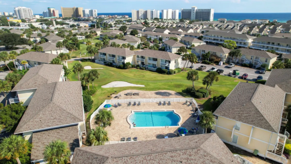 This vibrant exterior scene features a central swimming pool surrounded by lounge chairs, a well-maintained green golf putting area, rows of beach-style condominium buildings, and a distant view of high-rise hotels and the Gulf of Mexico. Activity is visible around the pool and golf green, with ample community space for recreation and relaxation.