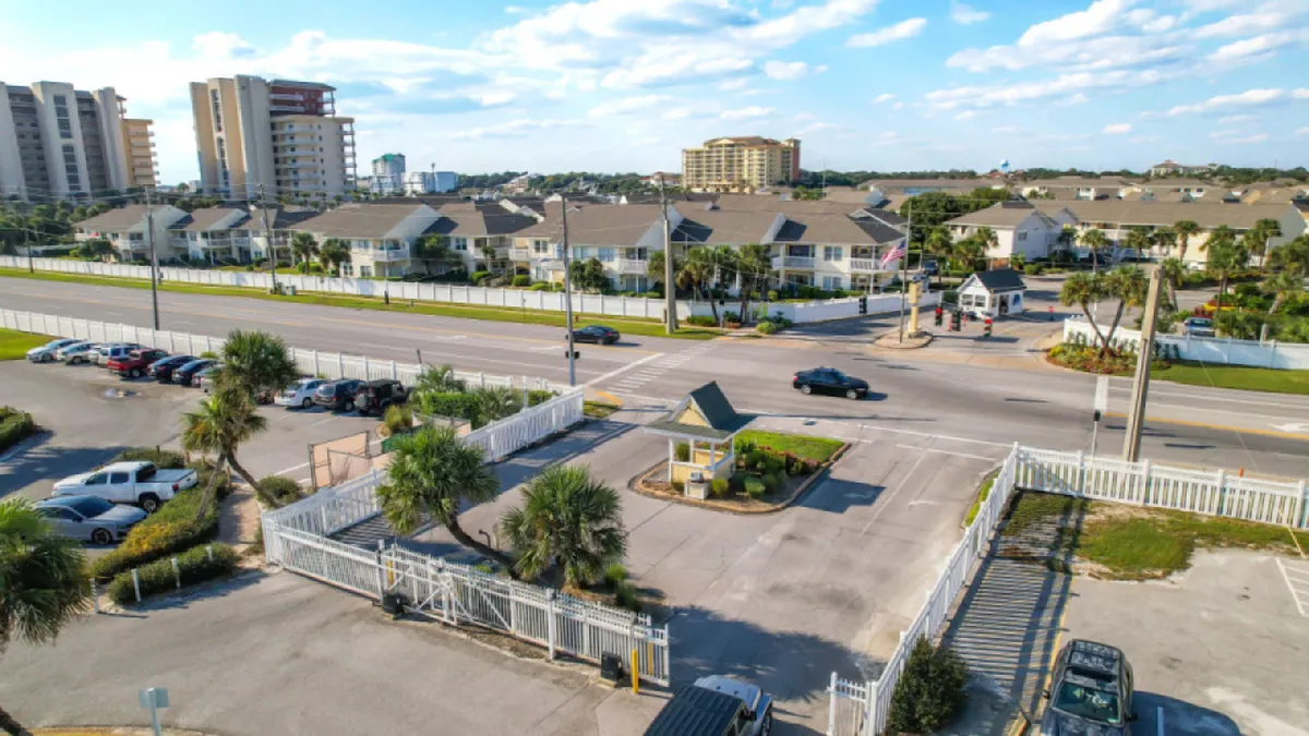 A welcoming gated entrance with a guardhouse marks the secured entry to the condo community. White fencing surrounds the property, and a parking lot with various cars can be seen. In the distance, residential and hotel buildings hint at the proximity to both home comforts and resort amenities.