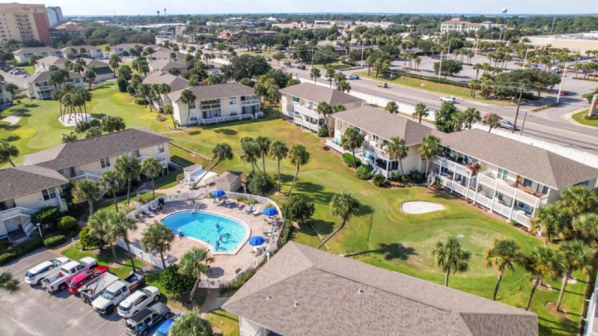 A vibrant exterior view showcasing a central swimming pool with lounge chairs and umbrellas surrounded by two-story condominium buildings. In the foreground, there's a putting green area for pitch and putt golf, and the landscaped grounds create a welcoming, family-friendly resort atmosphere.