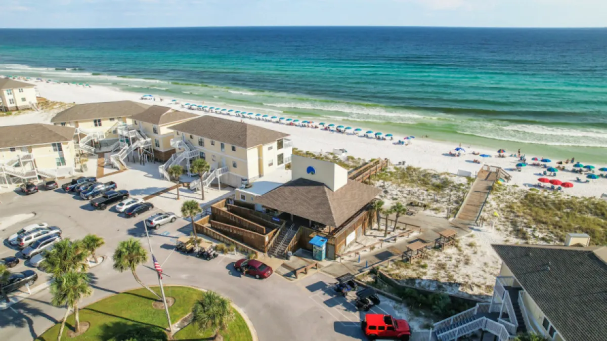 A vibrant stretch of white sandy beach is dotted with colorful umbrellas and lively visitors enjoying the turquoise Gulf waters. Beachfront condos and a boardwalk are visible, suggesting both the accessibility and the lively, communal atmosphere of the Emerald Coast.