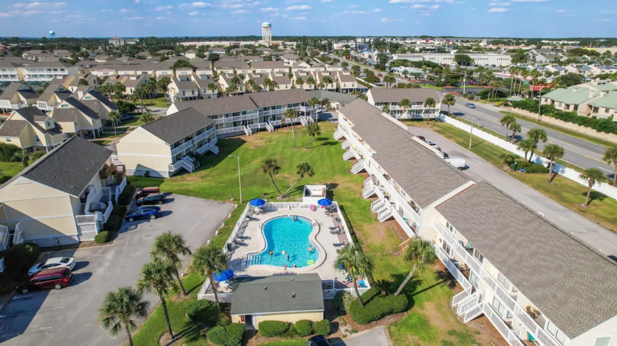 A central swimming pool is surrounded by lounge chairs and umbrellas, with palm trees and green lawns adding to the inviting, vacation atmosphere. Multi-unit condo buildings surround the pool, and a parking area sits nearby, showcasing the community's layout and amenities.