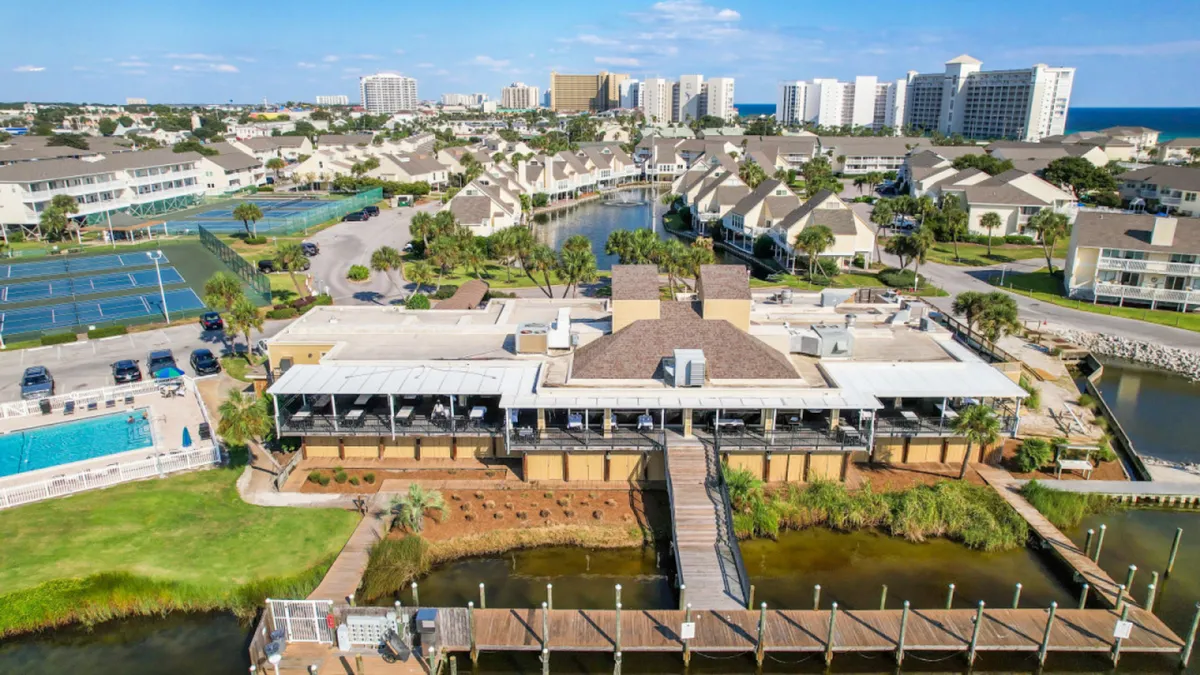 A comprehensive exterior scene including marina docks and waterfront, a community clubhouse or restaurant with a covered patio, visible tennis and pickleball courts, and a swimming pool area. The scene is lively, inviting, and hints at the breadth of activities and amenities within the resort.